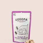 A stand-up pouch of GOGOMA freeze-dried pet treats on a light beige background, with several bite-sized, cube-shaped treats placed in front of the package. The package is white with a purple border. black text, and  illustration of a deer and sweet potato.