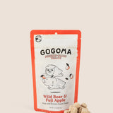 A stand-up pouch of GOGOMA freeze-dried pet treats on a warm beige background, with several small cube-shaped treat bites placed in front of the package. The package has a red border and a white center with black text reading GOGOMA at top of pouch and red text reading Wild Boar & Fuji Apple on bottom of pouch and an illustration of a boar and sweet potato.
