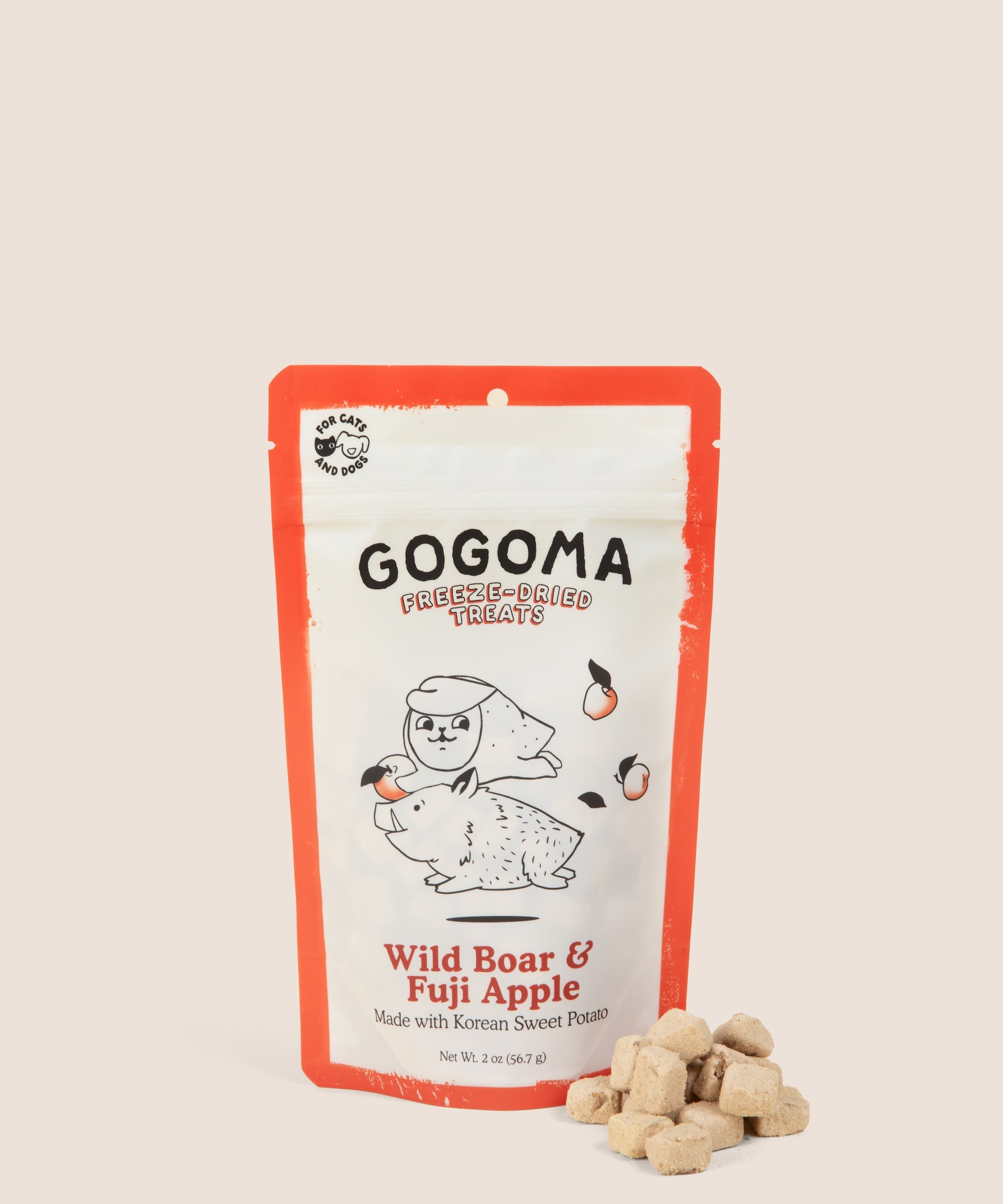 A stand-up pouch of GOGOMA freeze-dried pet treats on a warm beige background, with several small cube-shaped treat bites placed in front of the package. The package has a red border and a white center with black text reading GOGOMA at top of pouch and red text reading Wild Boar & Fuji Apple on bottom of pouch and an illustration of a boar and sweet potato.