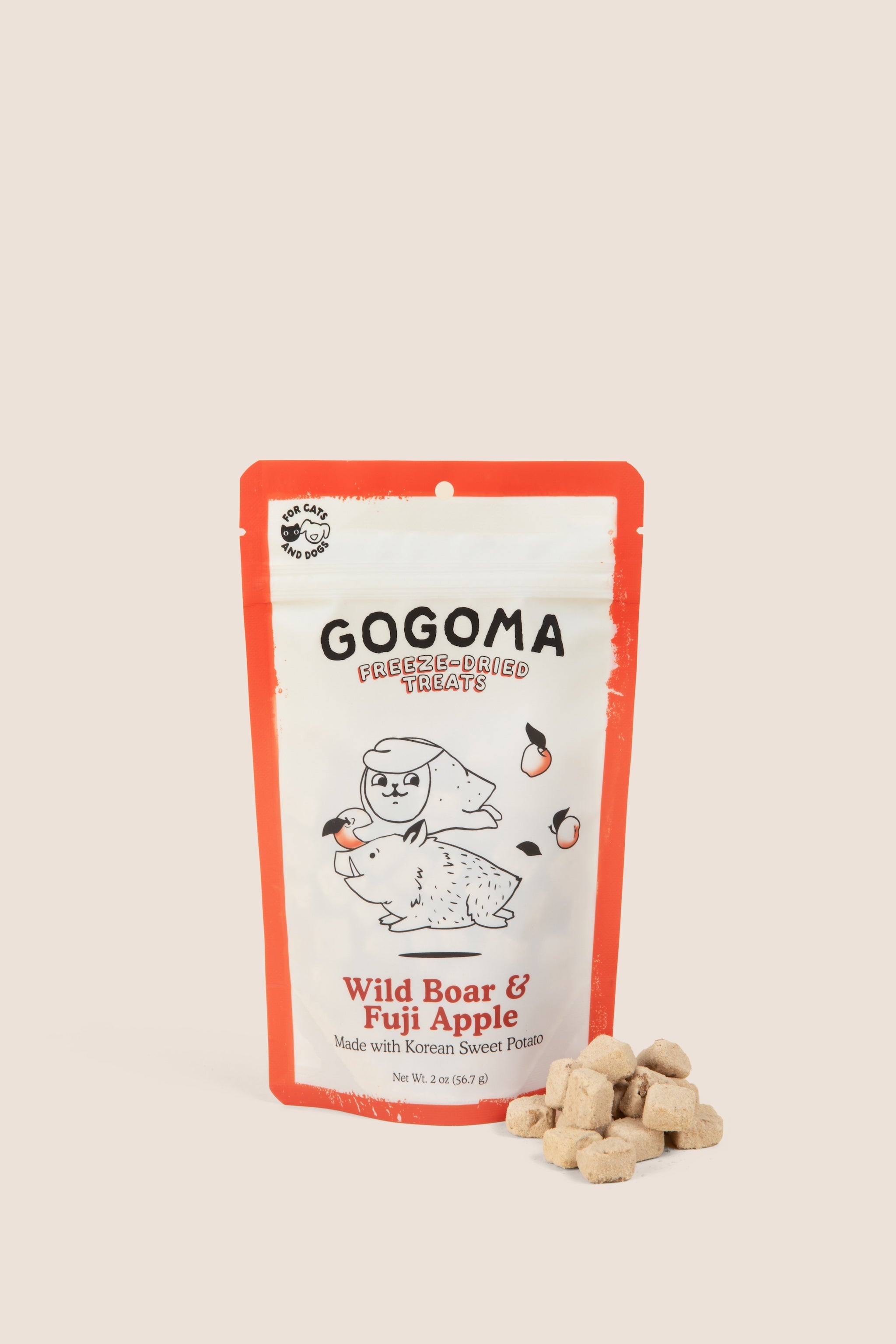 A stand-up pouch of GOGOMA freeze-dried pet treats on a warm beige background, with several small cube-shaped treat bites placed in front of the package. The package has a red border and a white center with black text reading GOGOMA at top of pouch and red text reading Wild Boar & Fuji Apple on bottom of pouch and an illustration of a boar and sweet potato.